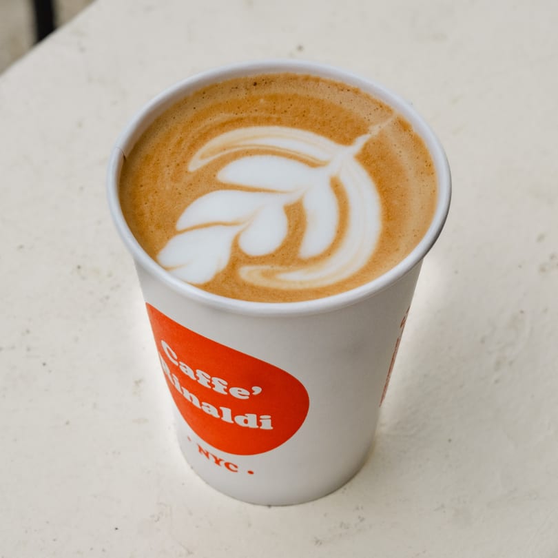 A Caffe' Rinaldi NYC branded cup of latte with intricate leaf art on top.