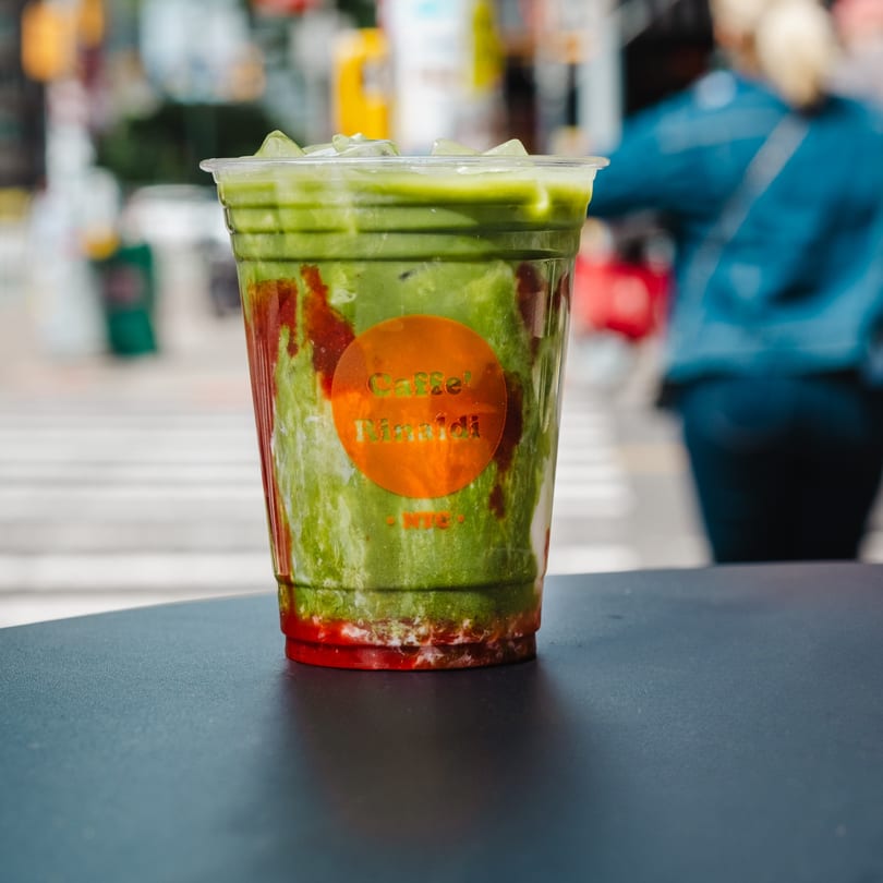 Iced matcha latte with strawberry syrup and ice, on a dark table with a blurred street background.