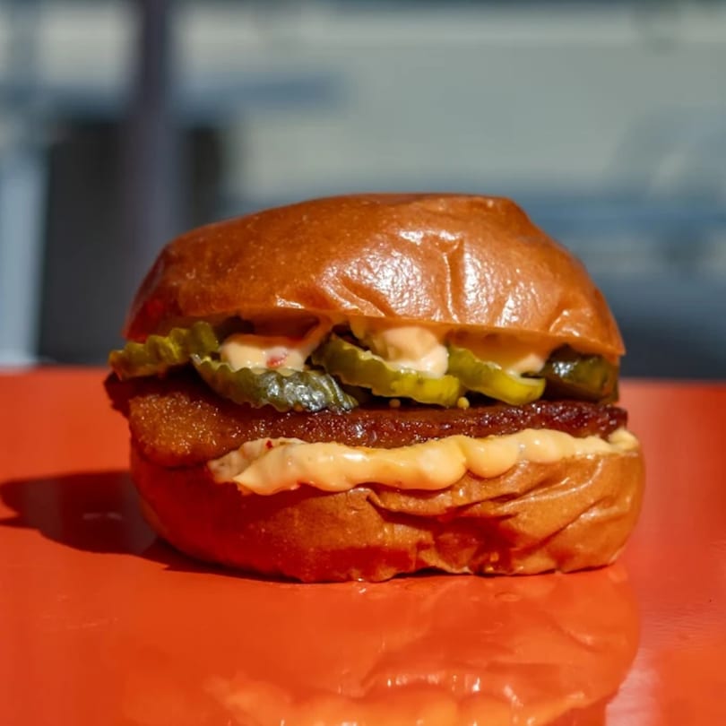 A close-up of a burger with pickles and sauce on a shiny orange surface.