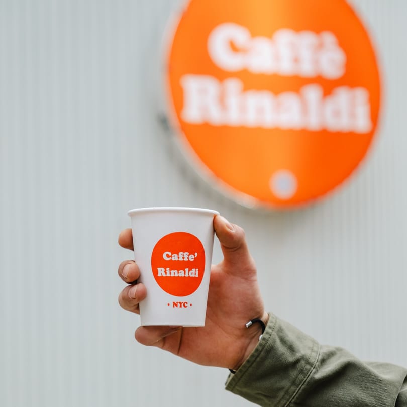 A hand holding a white Caffe' Rinaldi coffee cup in front of a blurred orange sign.