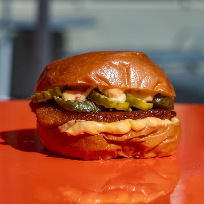 A close-up of a burger with pickles and sauce on a shiny orange surface.