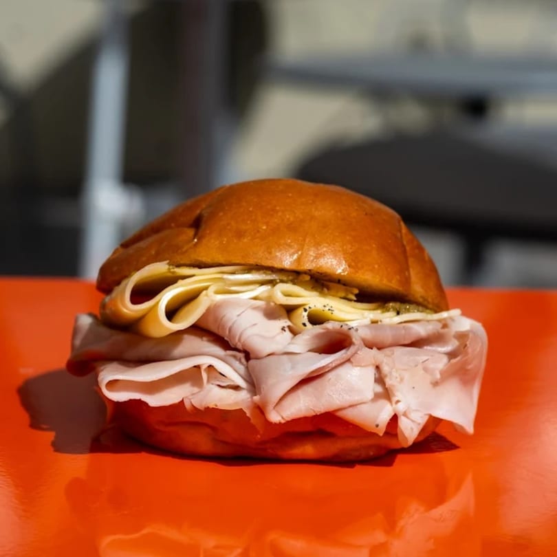 A large ham and cheese sandwich on a brioche bun sits on a bright orange surface.