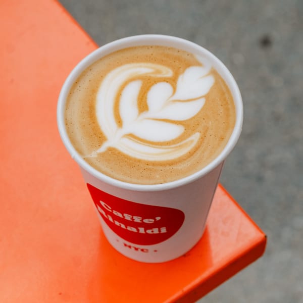 A Caffe' Rinaldi NYC latte with leaf art sits on a bright orange table.
