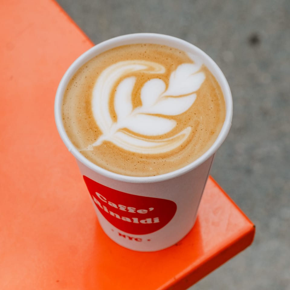 A Caffe' Rinaldi NYC latte with leaf art sits on a bright orange table.