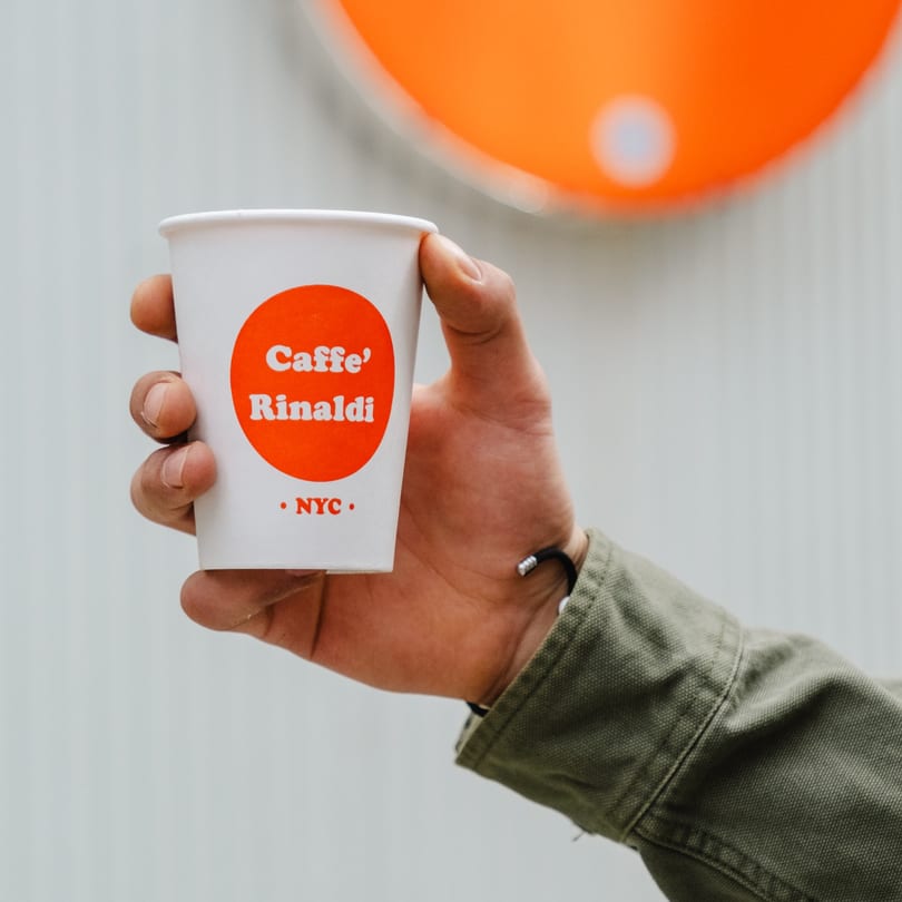 Hand holding a white Caffe' Rinaldi NYC coffee cup with an orange logo.