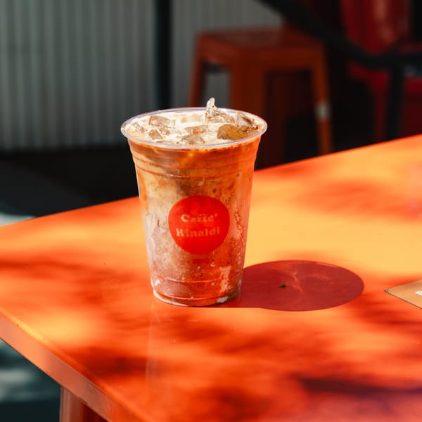Iced coffee with a red "Caffe' Rinaldi" sticker on an orange table in sunlight.