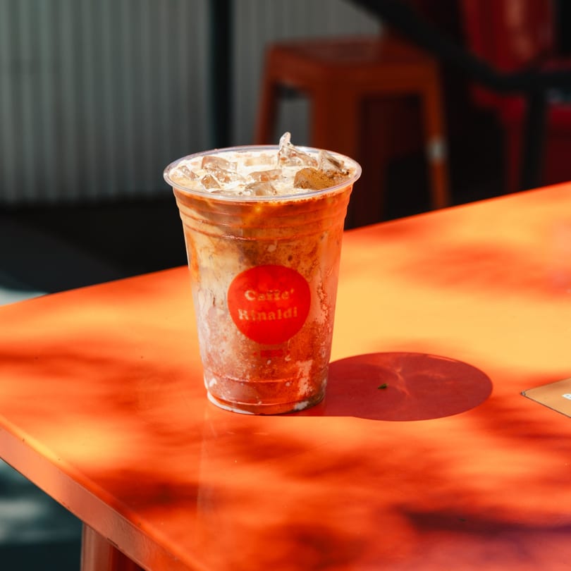 Iced coffee with a red "Caffe' Rinaldi" sticker on an orange table in sunlight.