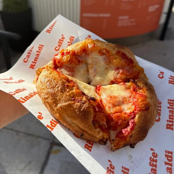 A hand holds a small, round pizza with melted cheese and tomato sauce on "Caffe' Rinaldi" branded paper.
