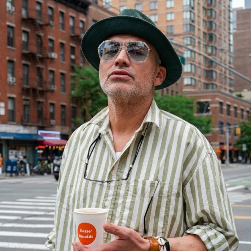 Man in a striped shirt and hat holding a "Caffè Rinaldi" cup on a city street.