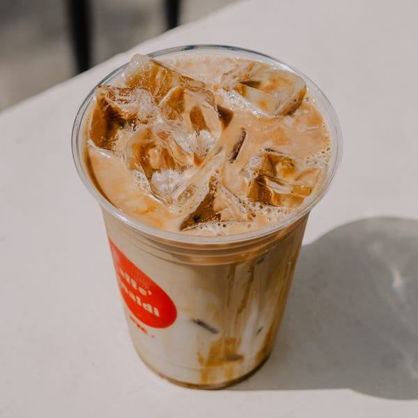Iced latte with a red logo on the cup, sitting on a white table in sunlight.
