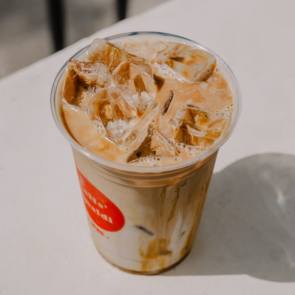 Iced latte with a red logo on the cup, sitting on a white table in sunlight.