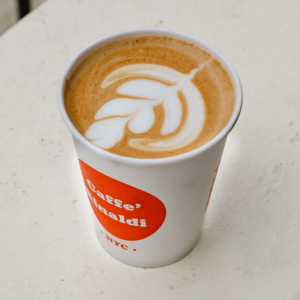 A Caffe' Rinaldi NYC branded cup of latte with intricate leaf art on top.