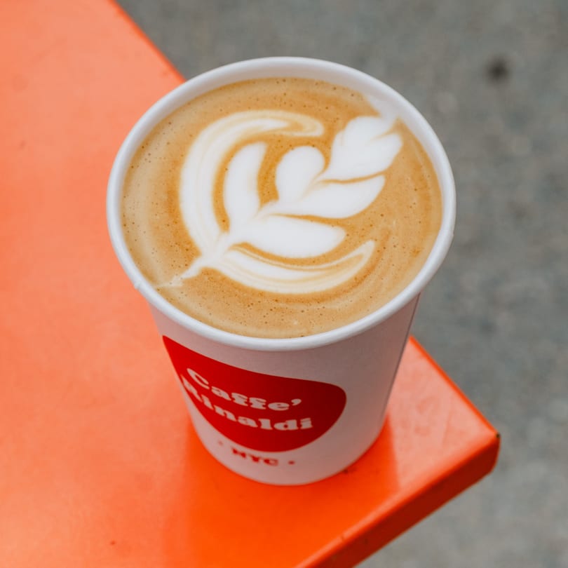 A Caffe' Rinaldi NYC latte with leaf art sits on a bright orange table.