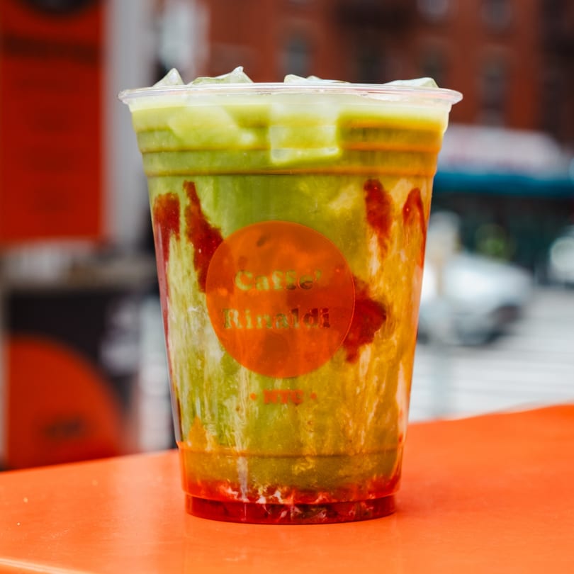 Iced matcha latte with strawberry syrup and a "Caffe' Rinaldi" logo on an orange table.