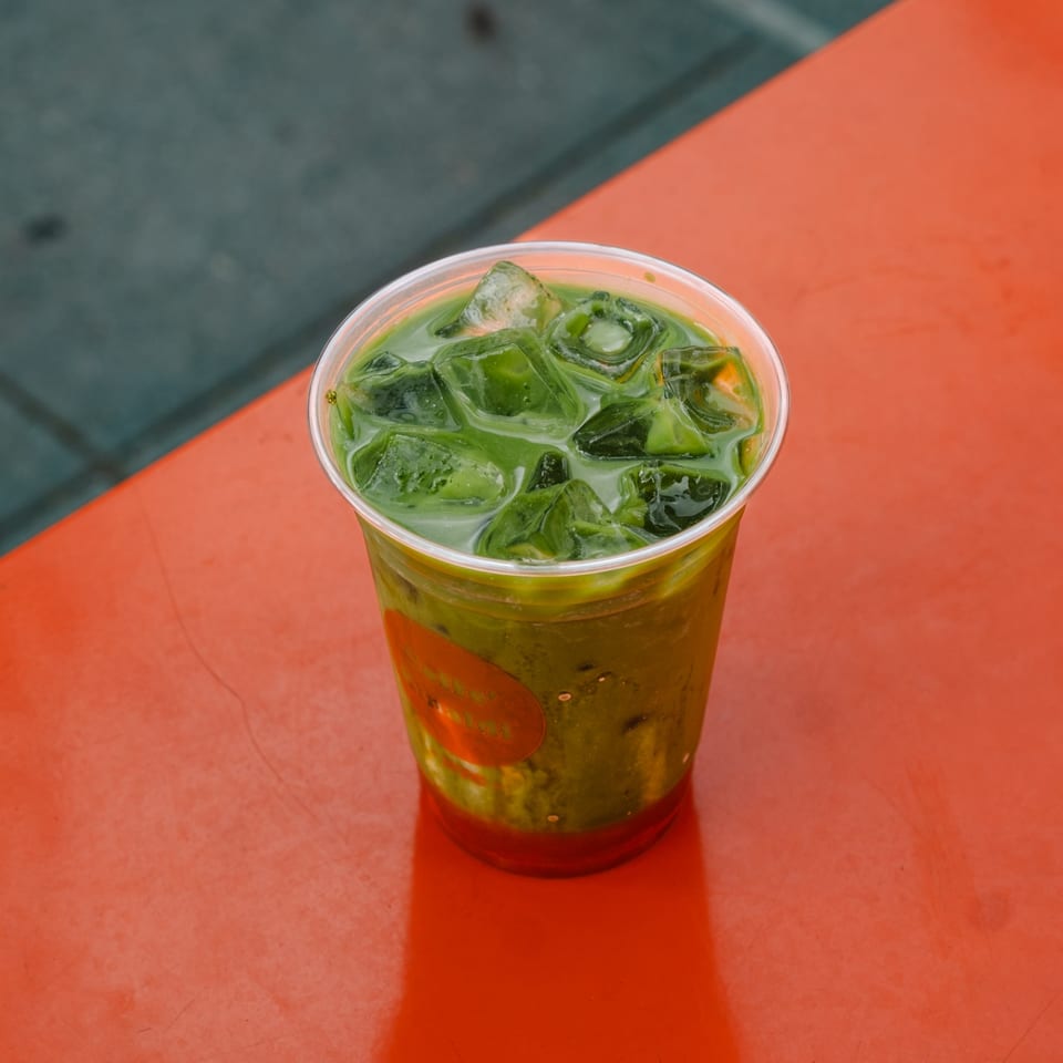 Iced green matcha drink with ice cubes in a clear plastic cup on a vibrant orange table.